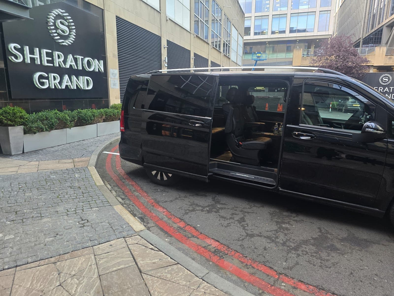 Black Mercedes-Benz V-Class with its sliding door open, ready for passengers outside a hotel